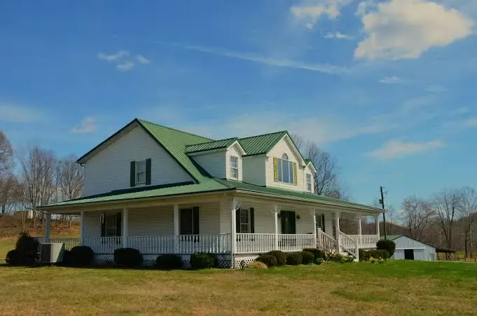 White farmhouse with green metal roof, wrap-around porch, green shutters, and open yard showing classic rural curb appeal