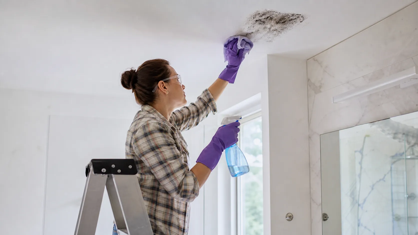 How to remove mold from painted ceiling: woman wearing gloves and safety glasses cleaning mold with a cloth and spray bottle from a bathroom ceiling