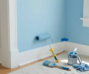 Do you paint trim or walls first guide showing freshly painted white baseboard trim next to a light blue wall, with paint roller, angled brush, and paint can on a drop cloth