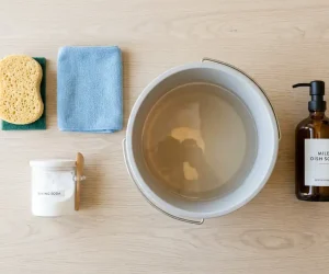 Flat-lay of wall cleaning supplies including sponge, microfiber cloth, baking soda, warm water bucket, and mild dish soap for how to clean painted walls safely