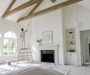 How to paint cathedral ceilings: a painter on a platform ladder rolls white paint on a vaulted ceiling with wood beams using an extension pole.