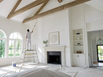 How to paint cathedral ceilings: a painter on a platform ladder rolls white paint on a vaulted ceiling with wood beams using an extension pole.