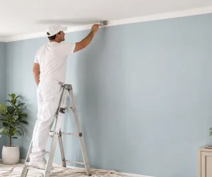 Professional painter on a ladder showing how to paint ceiling edges with an angled brush along a light blue wall and white ceiling