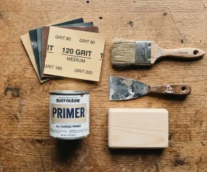 Sandpaper grits, Rust-Oleum primer, paintbrush, and putty knife laid out for how to prepare wood for painting