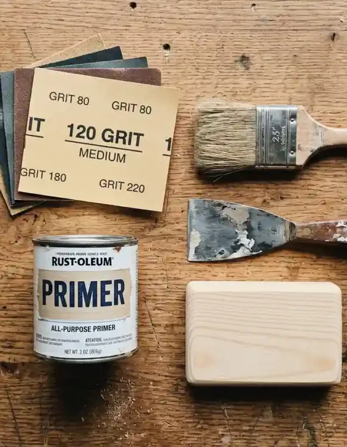 The basic tools for prepping wood before you paint: sandpaper grits, primer, a brush, and a scraper.