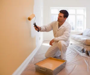 A professional painter wearing traditional white work clothes demonstrates proper interior painting technique. The white uniform helps conceal paint splatter and maintains a clean, professional appearance.